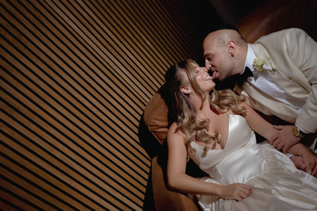 A Three-Day Destination Wedding in Tuscany 31 A bride and groom sit closely together, smiling and about to kiss. The bride wears a satin white gown, and the groom is in a white jacket with a black bow tie. They hold hands and share a joyful, intimate moment.