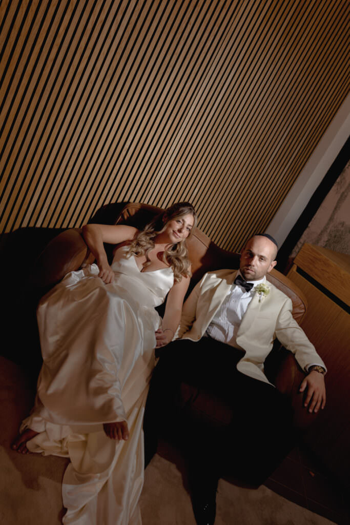 A Three-Day Destination Wedding in Tuscany 89 A bride in a white satin gown and a groom in a white tuxedo jacket sit relaxed on a brown sofa, holding hands, in a warmly lit room with wooden slat walls.