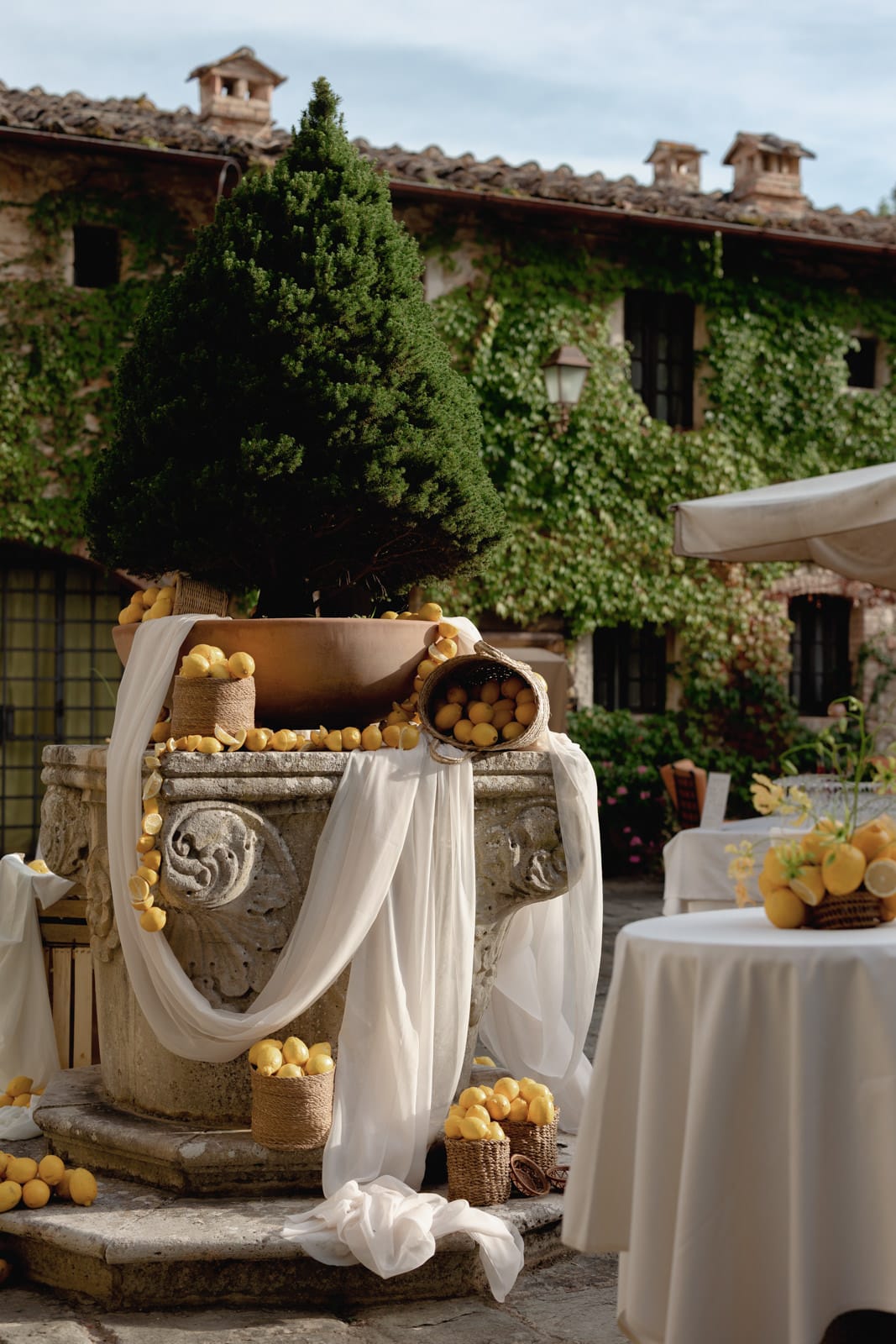 A rustic stone courtyard decorated with lemons, white fabric, and baskets. A large potted tree sits atop an ornate stone well, surrounded by lemons and baskets, with ivy-covered buildings in the background.