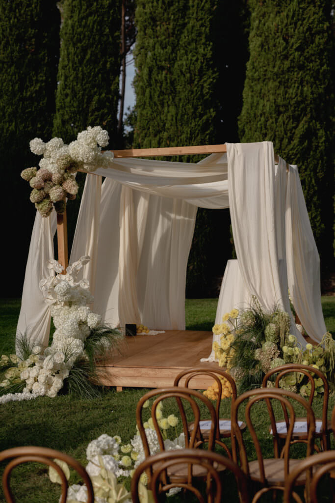 A Three-Day Destination Wedding in Tuscany 16 A wooden wedding arch draped with flowing cream fabric is decorated with white and pale yellow flowers, set outdoors on grass with tall green trees in the background and wooden chairs in the foreground.
