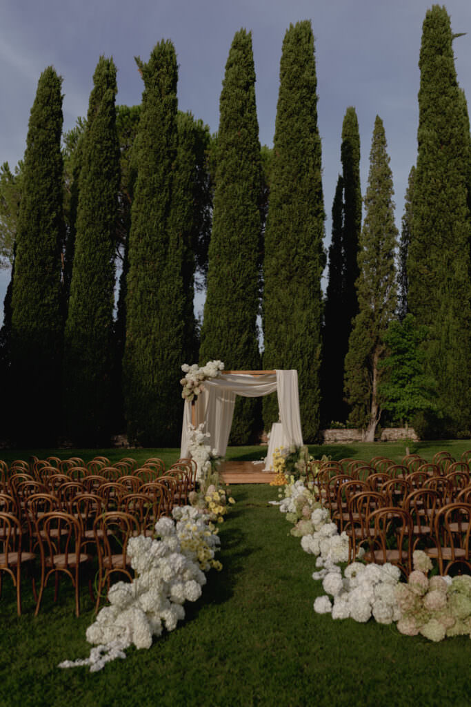 A Three-Day Destination Wedding in Tuscany 72 An outdoor wedding setup with a flower-lined aisle, wooden chairs, and a white draped arch beneath tall cypress trees on a lush green lawn.
