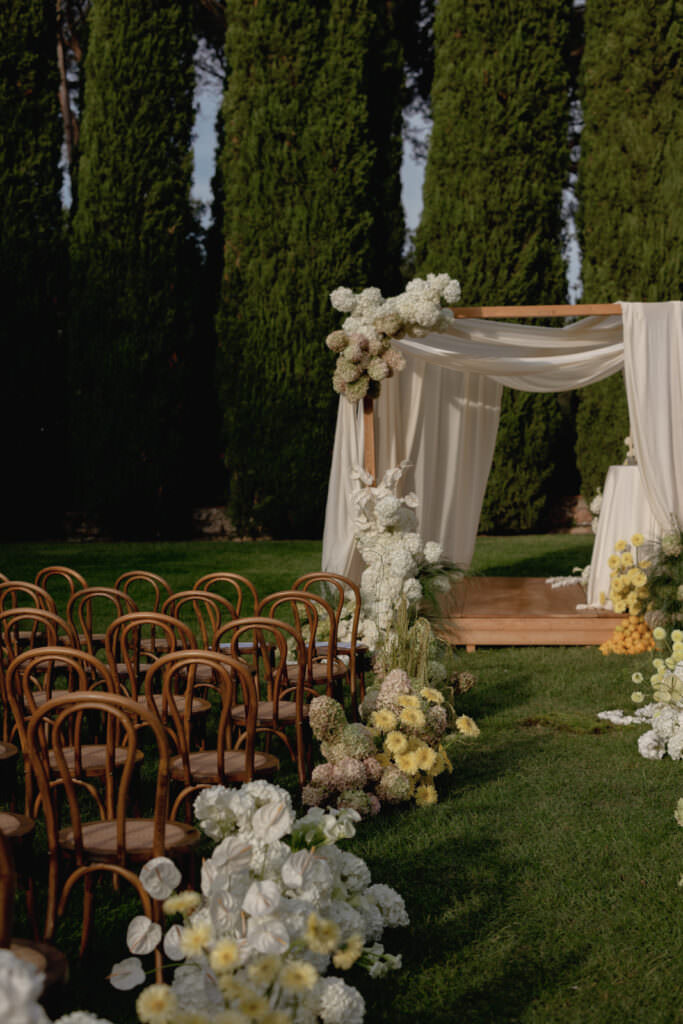 A Three-Day Destination Wedding in Tuscany 60 An outdoor wedding setup with wooden chairs in rows on grass, facing a wooden arch draped in white fabric and decorated with white and yellow flowers, with tall green trees in the background.