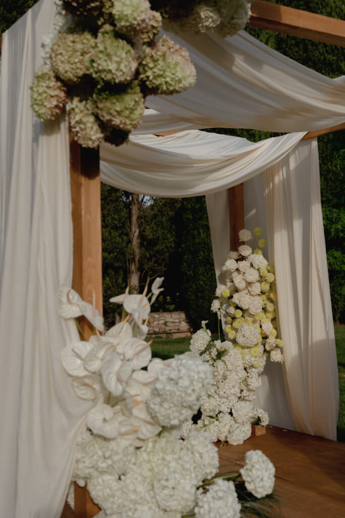 A Three-Day Destination Wedding in Tuscany 19 A wooden wedding arch draped with white fabric and decorated with clusters of white and light green flowers, set outdoors with greenery in the background.