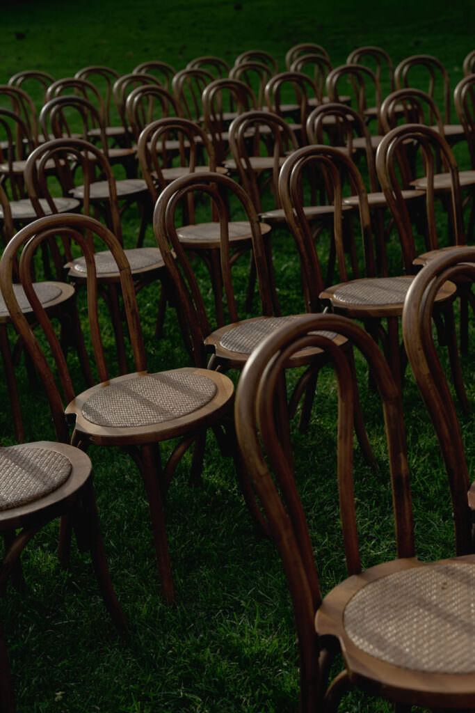 A Three-Day Destination Wedding in Tuscany 64 Rows of empty wooden chairs with curved backs are arranged outdoors on green grass, suggesting a setup for an event or gathering. The lighting creates shadows, adding depth to the scene.