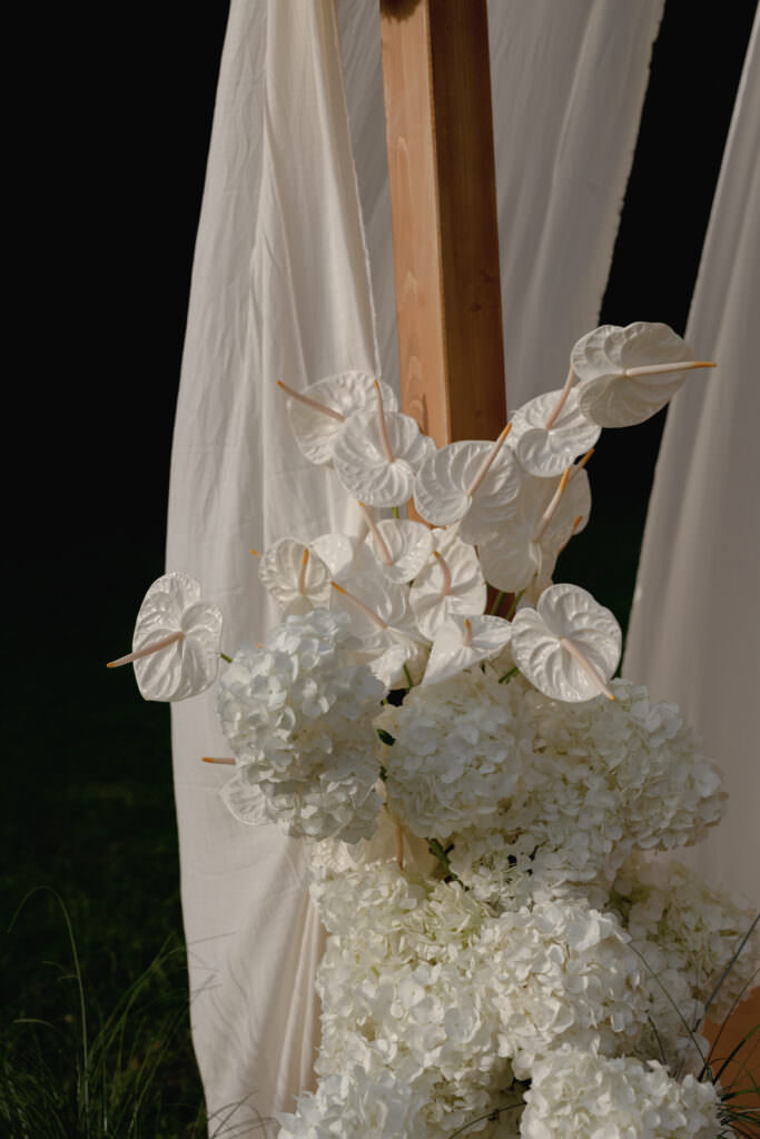 A Three-Day Destination Wedding in Tuscany 91 A wooden post is draped with white fabric and decorated with clusters of white hydrangeas and white anthurium flowers, set against a dark outdoor background.