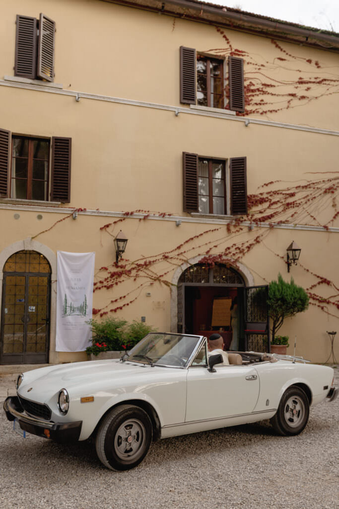 A Three-Day Destination Wedding in Tuscany 7 A vintage white convertible car is parked in front of a yellow building with brown shutters, climbing vines, and a banner near the entrance. The scene has a classic, European ambiance.