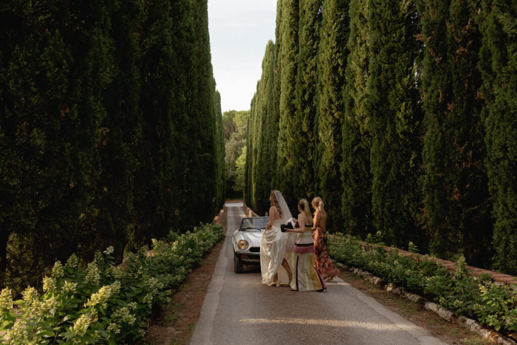 A Three-Day Destination Wedding in Tuscany 37 A bride in a white gown stands beside a vintage convertible on a tree-lined road, accompanied by two women in colorful dresses. Tall, dense green cypress trees rise on both sides.