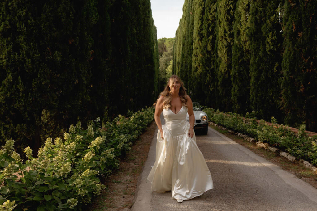 A Three-Day Destination Wedding in Tuscany 6 A woman in a white wedding dress walks on a tree-lined road, with a classic car parked behind her and tall green hedges on both sides.