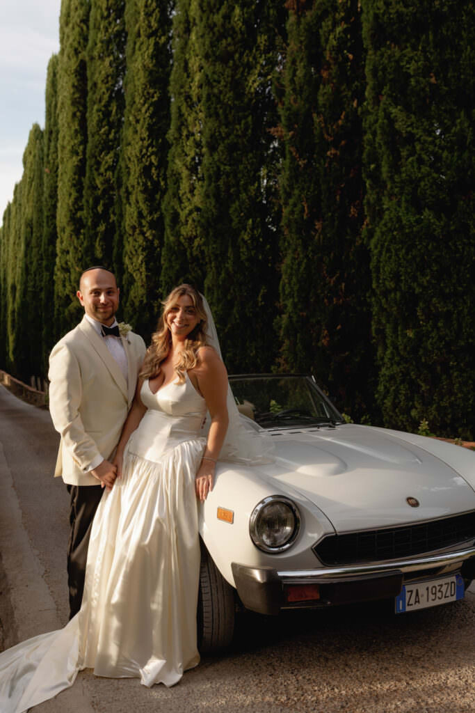 A Three-Day Destination Wedding in Tuscany 70 A bride and groom in formal wedding attire stand next to a classic white convertible car, parked on a road lined with tall green cypress trees. The bride leans on the car and both are smiling at the camera.