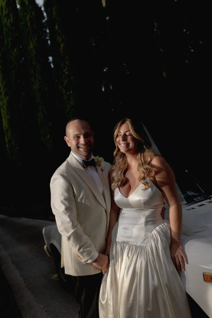 A Three-Day Destination Wedding in Tuscany 8 A smiling couple stands together outdoors by a white car. The man wears a white tuxedo jacket and bow tie, while the woman wears a white wedding gown with a fitted bodice and long, wavy hair. They appear happy and relaxed.