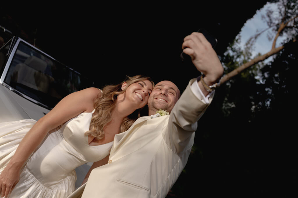 A Three-Day Destination Wedding in Tuscany 15 A joyful bride and groom in white wedding attire smile and lean together as they take a selfie outdoors, with trees and a white car visible in the background.