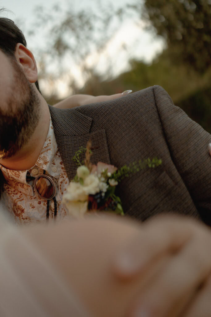 A close-up of a bearded man in a patterned shirt and brown jacket with a boutonnière, perfectly capturing the charm of an autumn wedding in a countryside style. Trees blur in the background as a hand rests gently on his shoulder.