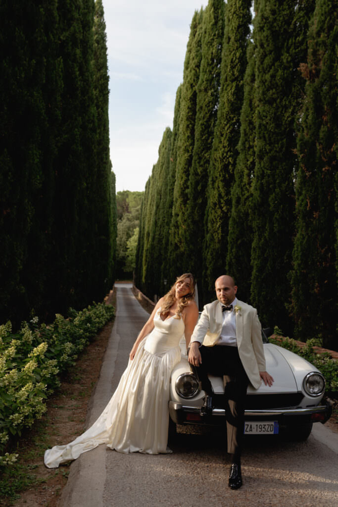 A Three-Day Destination Wedding in Tuscany 59 A bride and groom in formal attire pose by a classic white car on a narrow road lined with tall, dense green trees under a bright sky. The groom sits on the car while the bride stands beside him, both looking relaxed.