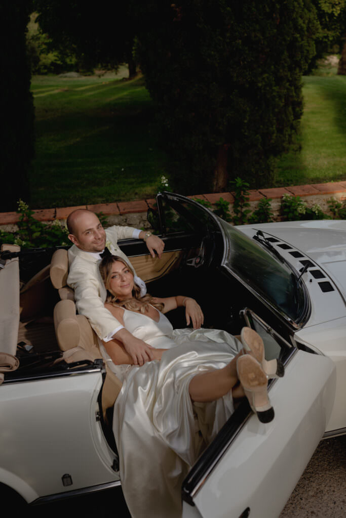 A Three-Day Destination Wedding in Tuscany 44 A couple dressed in elegant outfits relaxes in a white convertible car. The woman, in a satin dress, leans against the man, who has his arm around her. They appear happy, surrounded by greenery and sunlight.