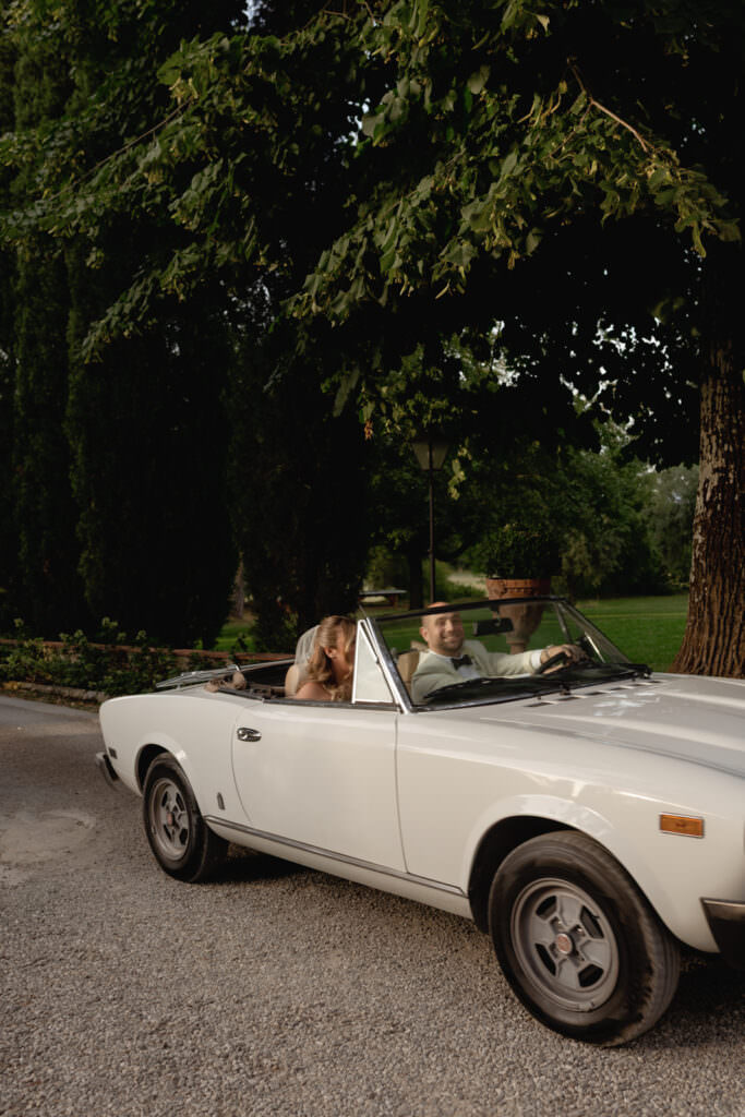 A Three-Day Destination Wedding in Tuscany 73 A couple sits in a vintage white convertible parked on a gravel path, surrounded by lush green trees and grass on a sunny day. The woman leans toward the man, both appearing happy and relaxed.