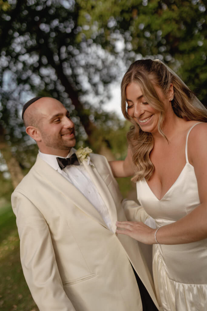 A Three-Day Destination Wedding in Tuscany 9 A bride in a satin gown and a groom in a cream tuxedo smile and look at each other outdoors, surrounded by greenery and sunlight, on their wedding day.