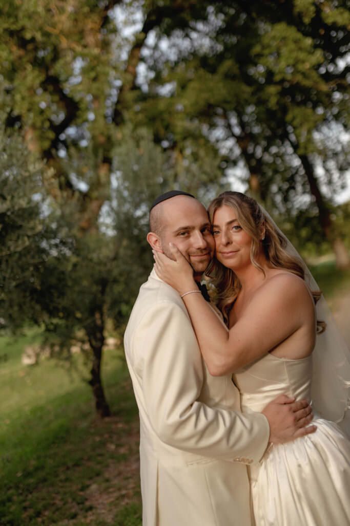 A Three-Day Destination Wedding in Tuscany 49 A bride and groom embrace outdoors, both wearing cream-colored wedding attire. They smile softly at the camera, surrounded by greenery and trees in a sunlit setting.