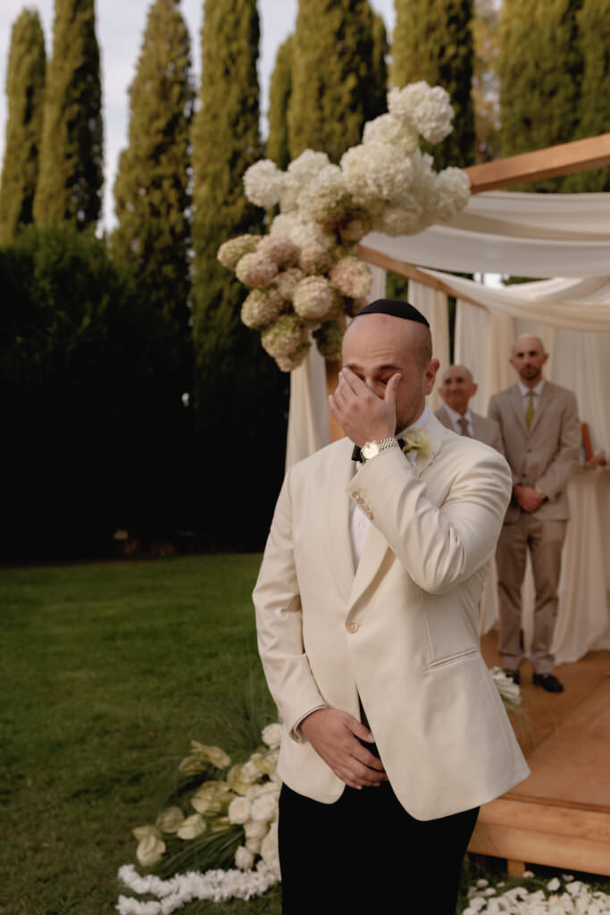 A Three-Day Destination Wedding in Tuscany 82 A groom in a cream-colored suit covers his face with his hand, appearing emotional, as he stands outdoors in front of a floral wedding arch with two men in the background.