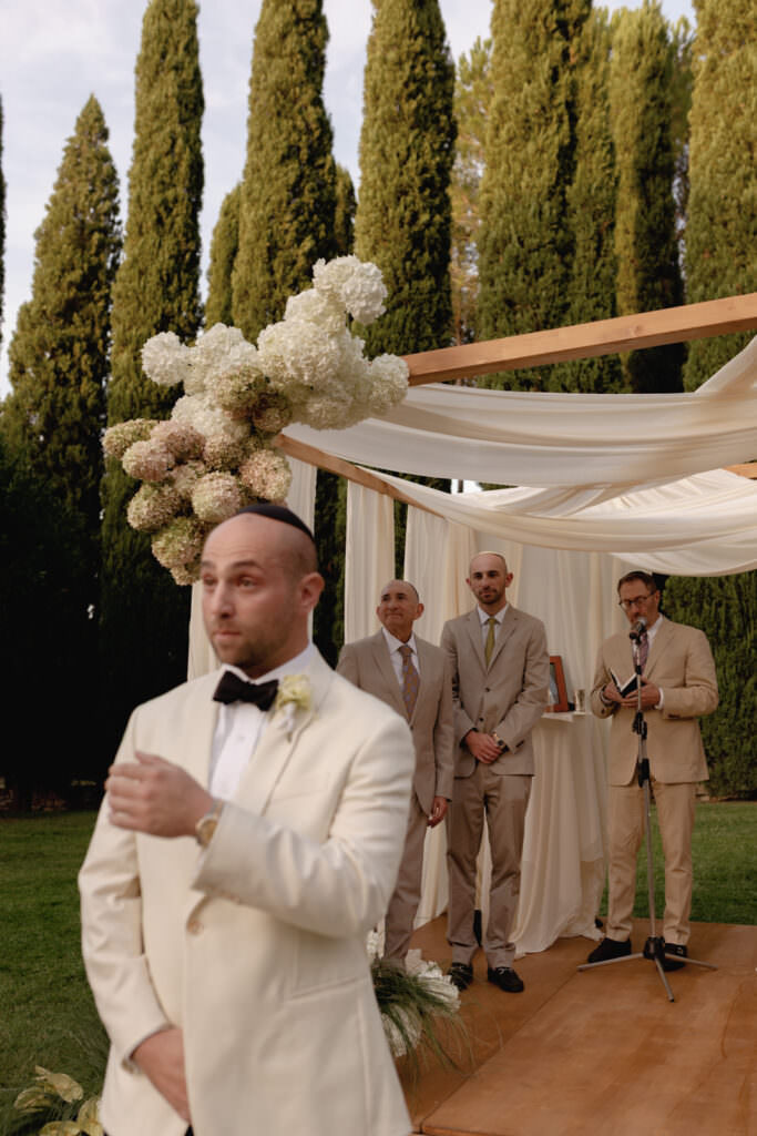 A Three-Day Destination Wedding in Tuscany 27 A man in a cream suit stands in the foreground, looking surprised or excited, while three men in beige suits stand under a floral-decorated wedding canopy outdoors, with tall trees in the background.