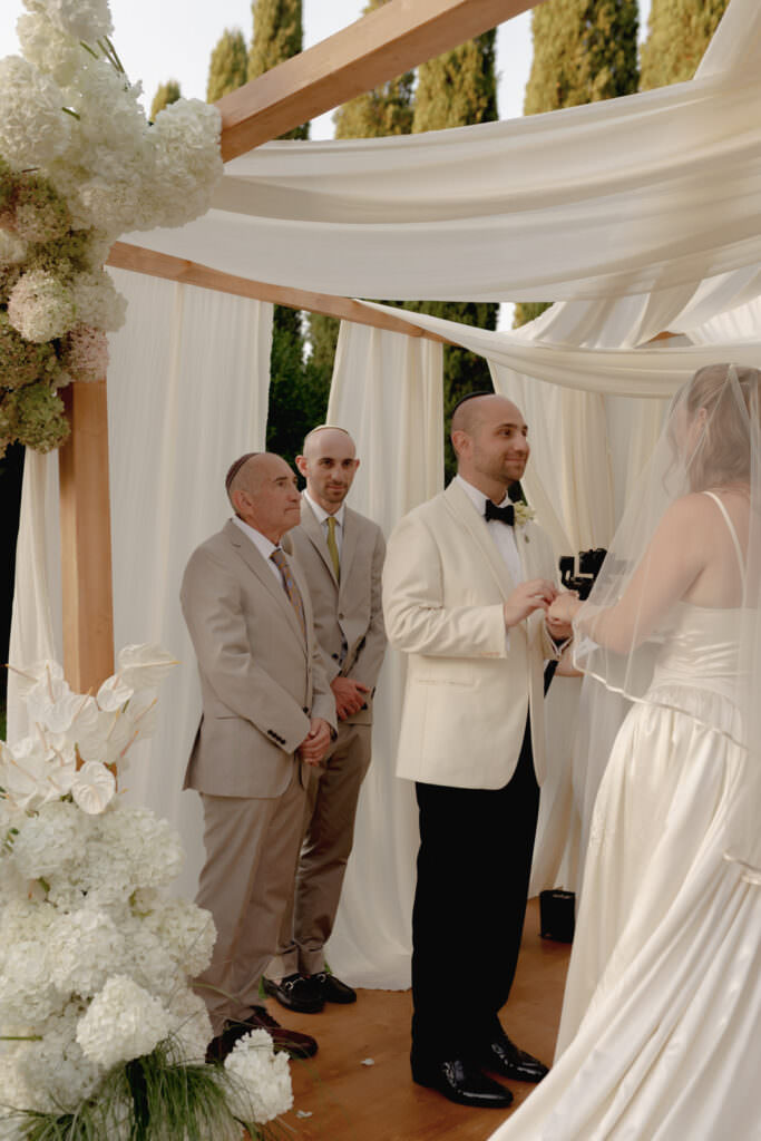 A Three-Day Destination Wedding in Tuscany 45 A bride and groom stand under a white, flower-decorated wedding canopy exchanging vows. The groom holds the brides hands, with two groomsmen in light suits standing behind him, and tall trees in the background.