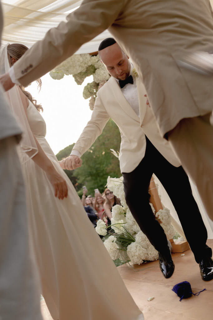 A Three-Day Destination Wedding in Tuscany 63 A groom in a white suit breaks a glass with his foot during a wedding ceremony under a floral canopy, while holding hands with the bride in a white dress. Guests watch and take photos in the background.