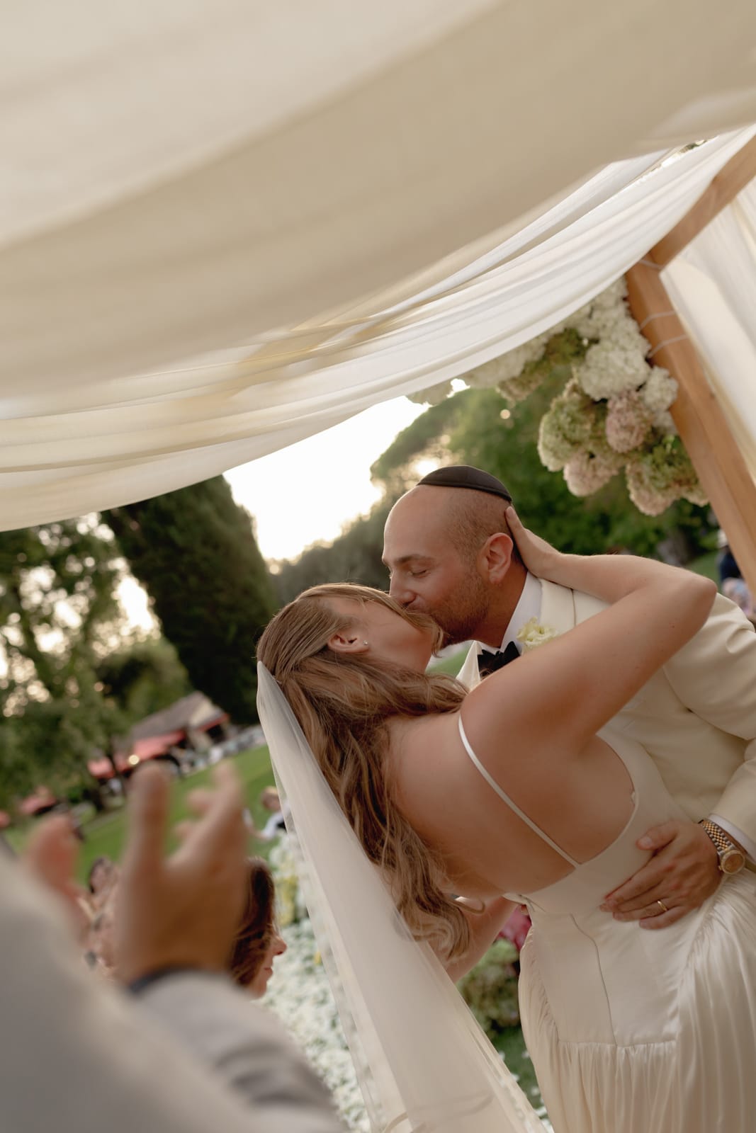 A bride and groom share a kiss under a floral wedding arch, surrounded by greenery. The bride wears a veil and white dress, while the groom wears a suit and yarmulke. Guests applaud in the foreground.