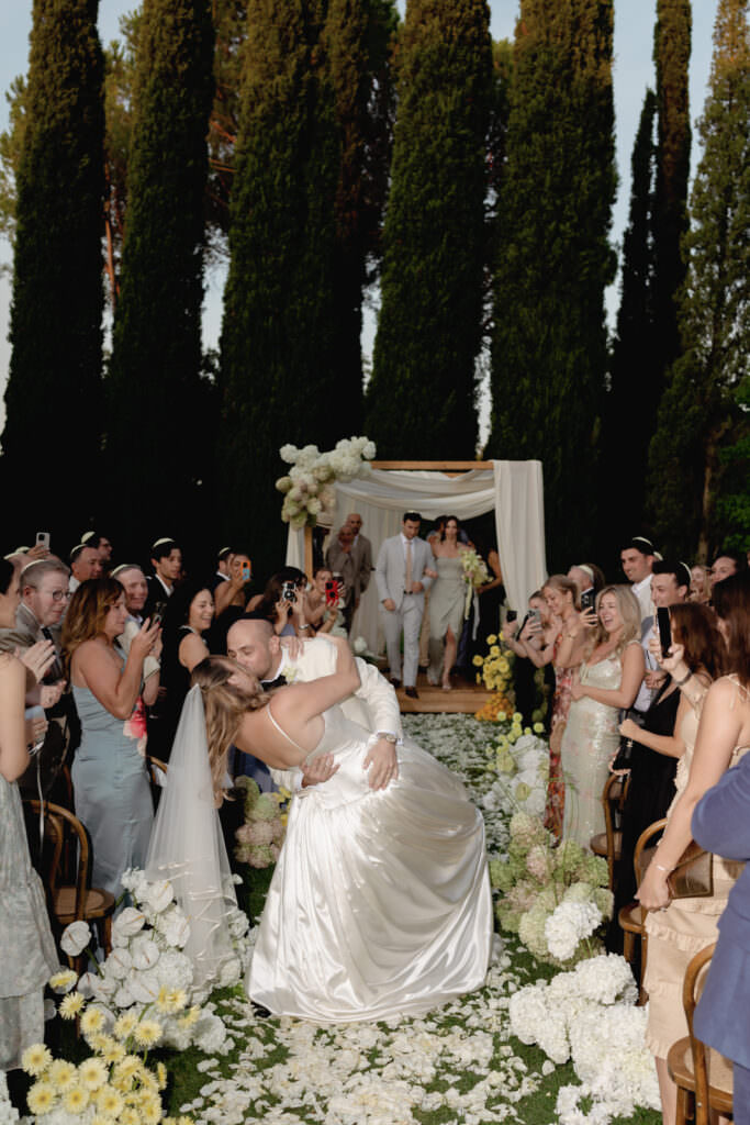 A Three-Day Destination Wedding in Tuscany 28 A bride and groom share a joyful dip and kiss in the aisle, surrounded by cheering guests and white flowers, with tall trees and a decorated wedding arch in the background.