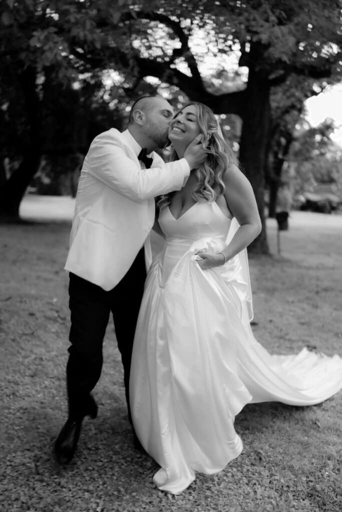 A Three-Day Destination Wedding in Tuscany 18 A groom in a white jacket kisses a smiling bride on the cheek as they walk outdoors under trees. The bride holds up her flowing white dress, and both look joyful and happy. The photo is in black and white.