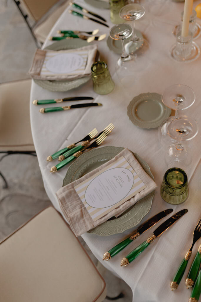 A Three-Day Destination Wedding in Tuscany 23 A round table set for a formal meal with green and gold cutlery, beige napkins, green glassware, elegant plates, and printed menus. Cream-colored chairs surround the table, which is covered with a white tablecloth.