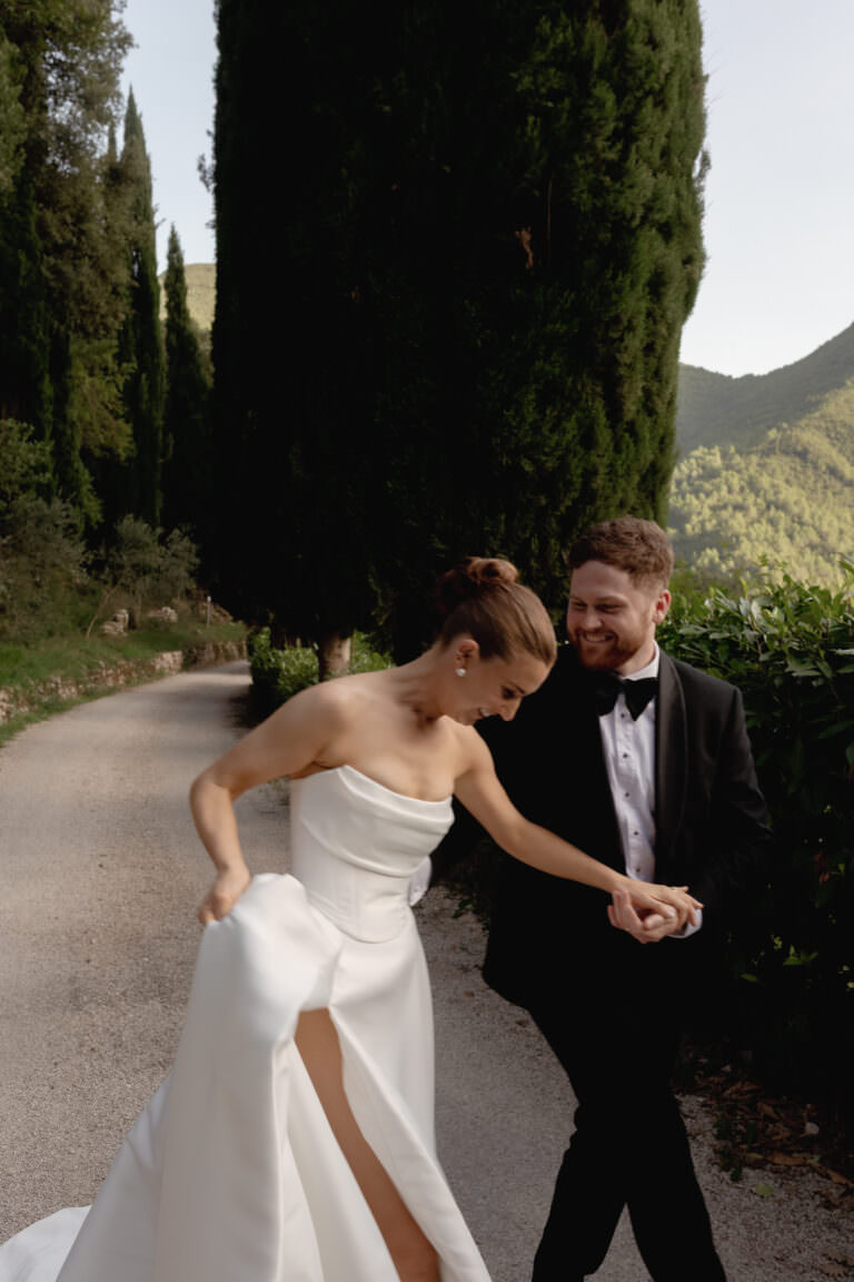Elegant Destination Wedding at San Pietro in Valle Abbey, Umbria 1 A bride in a white gown with a slit and a groom in a black tuxedo walk hand in hand on a path lined with tall trees, smiling joyfully at each other in a scenic outdoor setting.