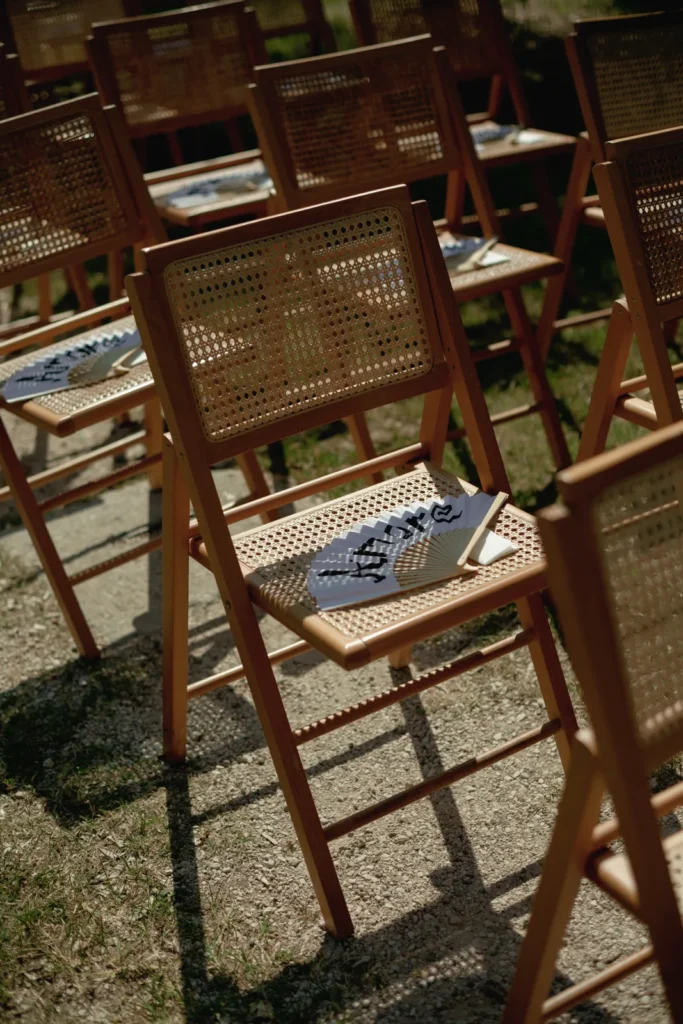 Home 42 Rows of wooden chairs with woven cane seats and backs are set up outdoors. Each chair has a folded program or booklet placed on the seat, and sunlight casts shadows on the ground.
