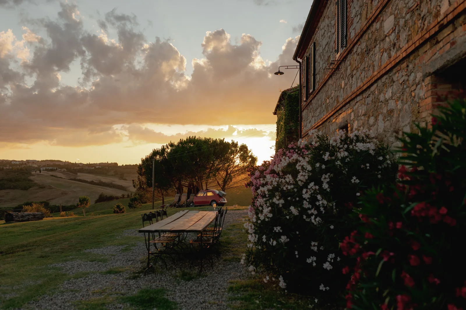A long wooden table and chairs sit on a grassy lawn beside a stone building with flowering bushes at sunset—an idyllic scene perfect for a Tuscan wedding photographer, overlooking rolling hills and a car parked near trees in the background.