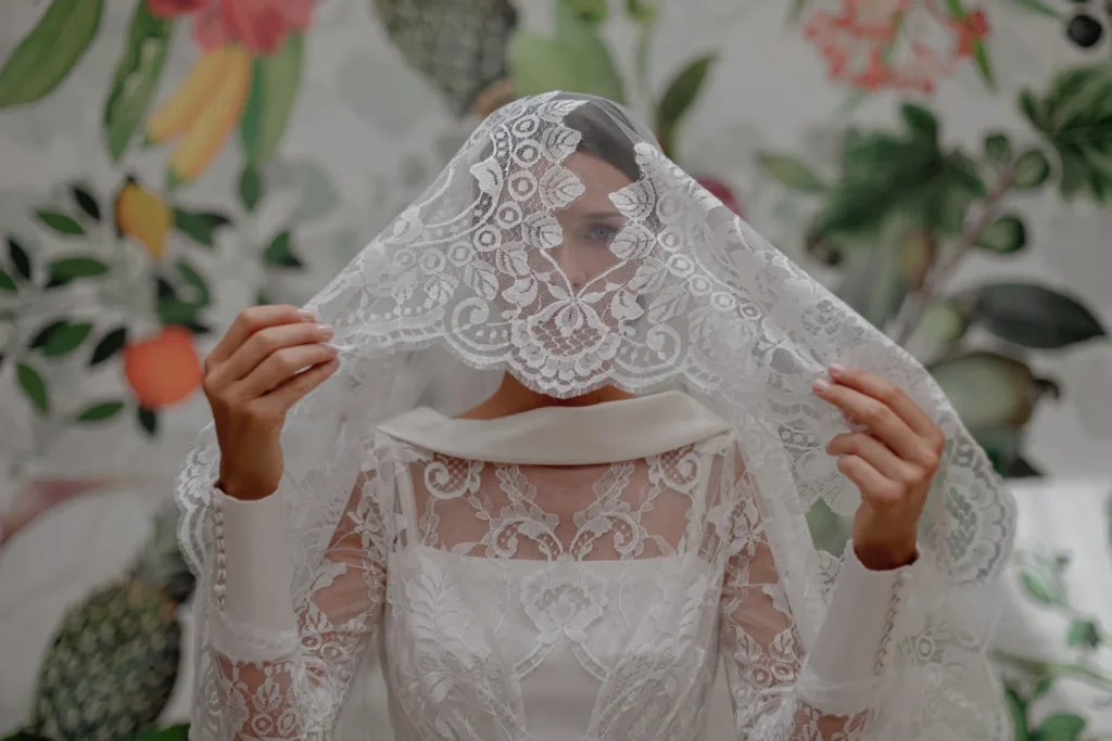 A bride in a white lace wedding dress holds a lace veil over her face, set against a colorful, floral and fruit-patterned backdrop—perfect for showcasing any destination wedding photographer portfolio.