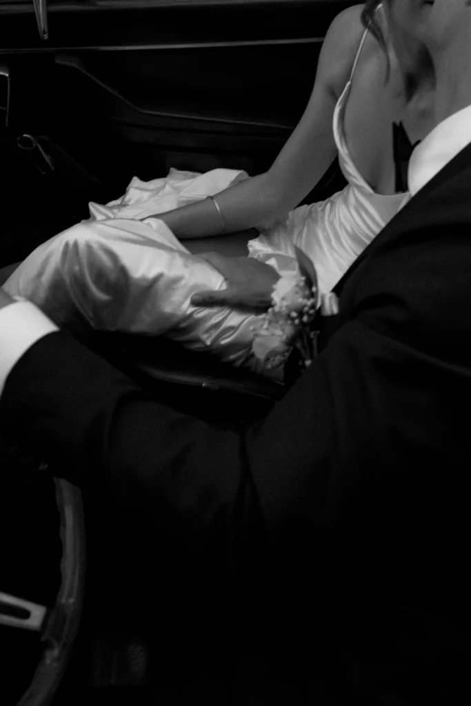 Black and white photo of a couple in formal attire sitting close together in a car; the woman wears a satin dress, and the man, in a suit, holds her leg gently while she rests her hand on his arm.