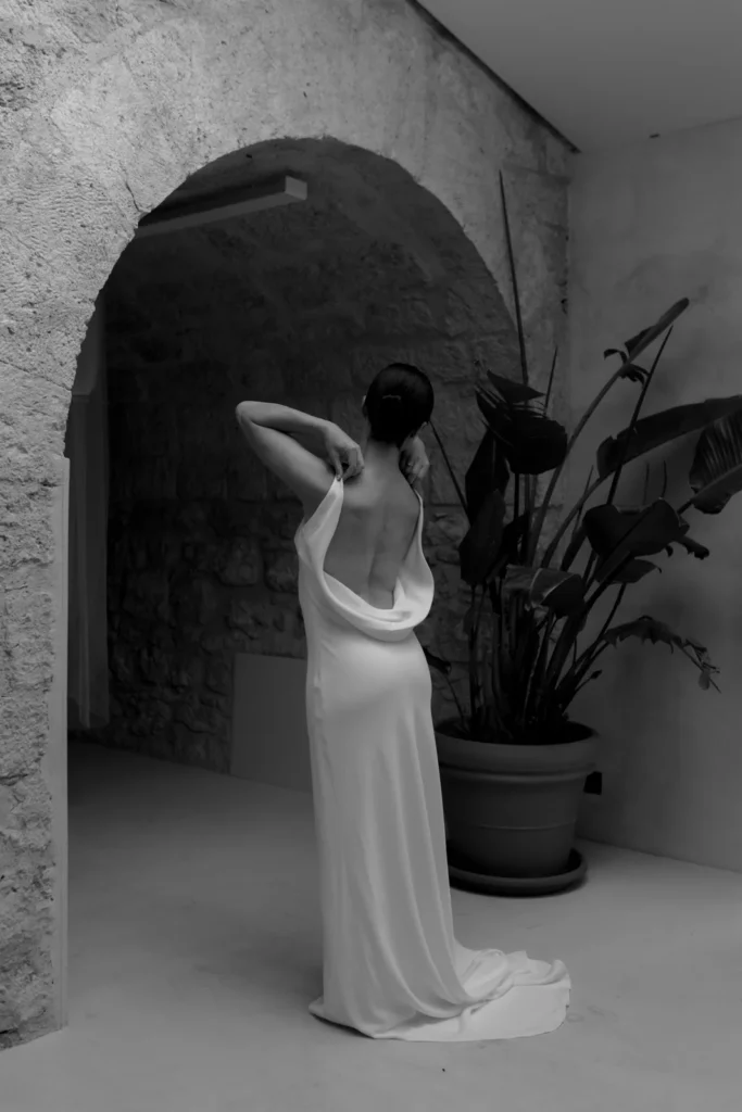 Home 43 A woman in a long, backless white dress stands indoors near a stone arch and large potted plant, facing away and adjusting her dress. The scene is in black and white.