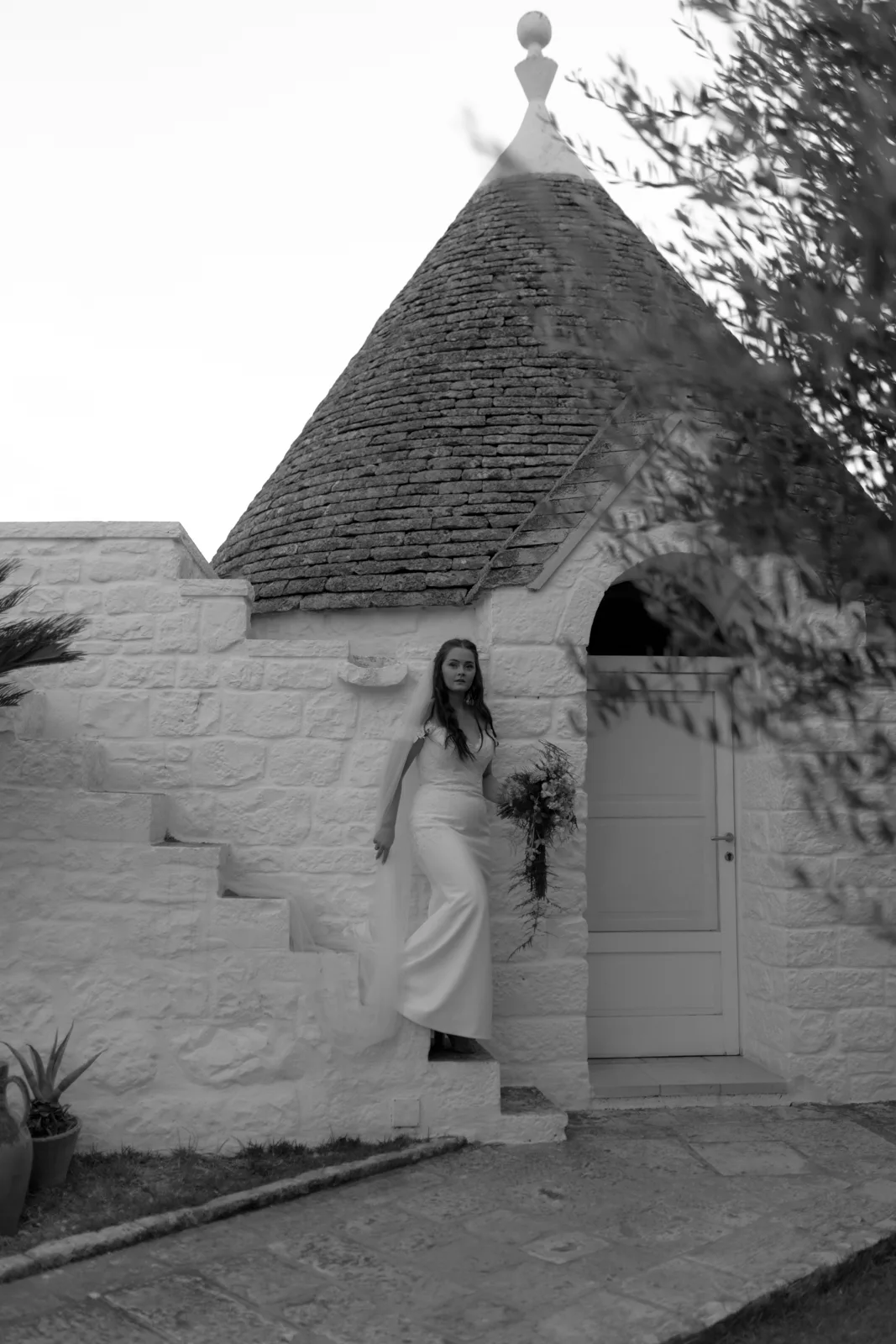 Home 38 A woman in a long dress holds a bouquet while standing on stone steps outside a white building with a conical roof. The scene is in black and white.