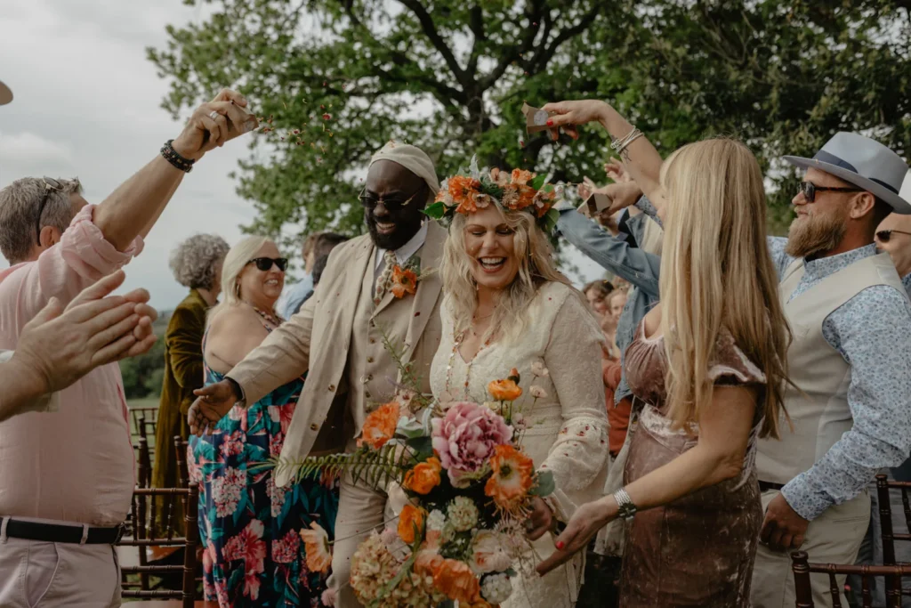 A joyful bride and groom walk down an outdoor aisle, smiling as guests cheer and toss flowers. Captured by an Italian wedding photographer, the bride holds a colorful bouquet and wears a floral crown; everyone looks happy and celebratory.