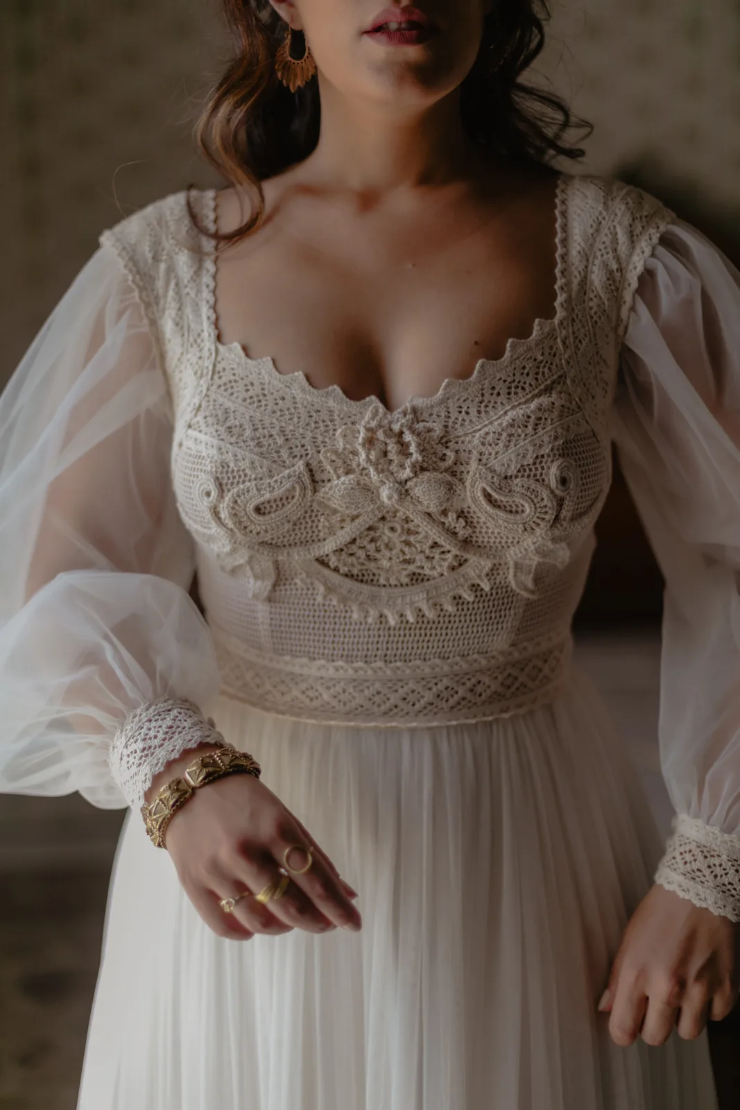Home 26 A woman wearing a vintage-style, lace embroidered white dress with sheer long sleeves and intricate crochet details, adorned with gold rings and a bracelet. Her face is partially out of frame.