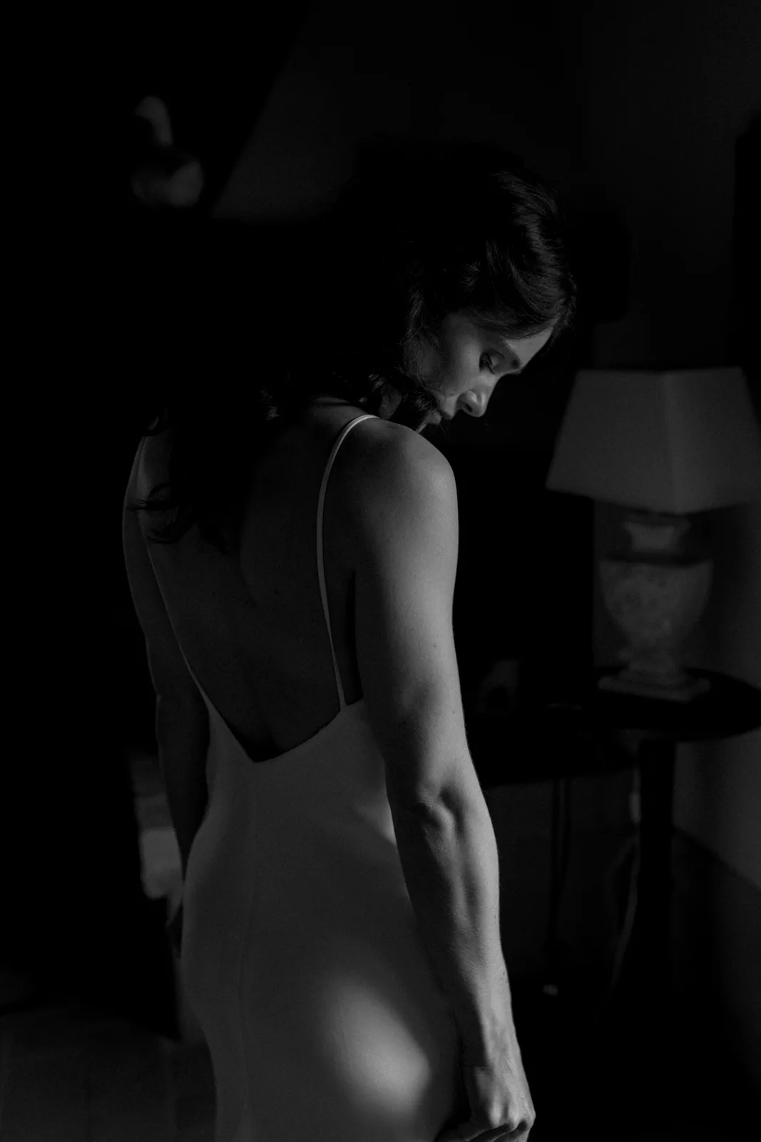 Home 23 A woman in a sleeveless dress stands in a dimly lit room, facing slightly away. Light highlights her bare back and arm, creating a dramatic shadow. A lamp and small table are visible in the background.