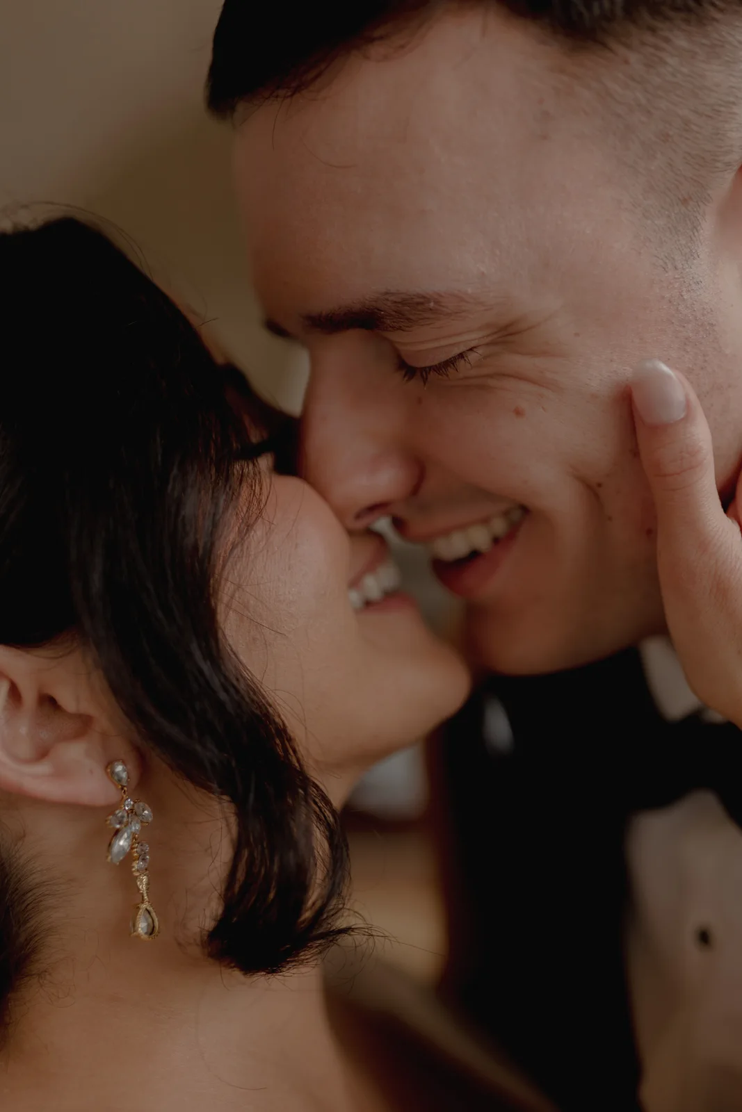 Kind Words 14 A close-up of a smiling couple about to kiss, with one person gently touching the others face and both appearing joyful. The woman wears a dangling earring and the man is dressed in formal attire.