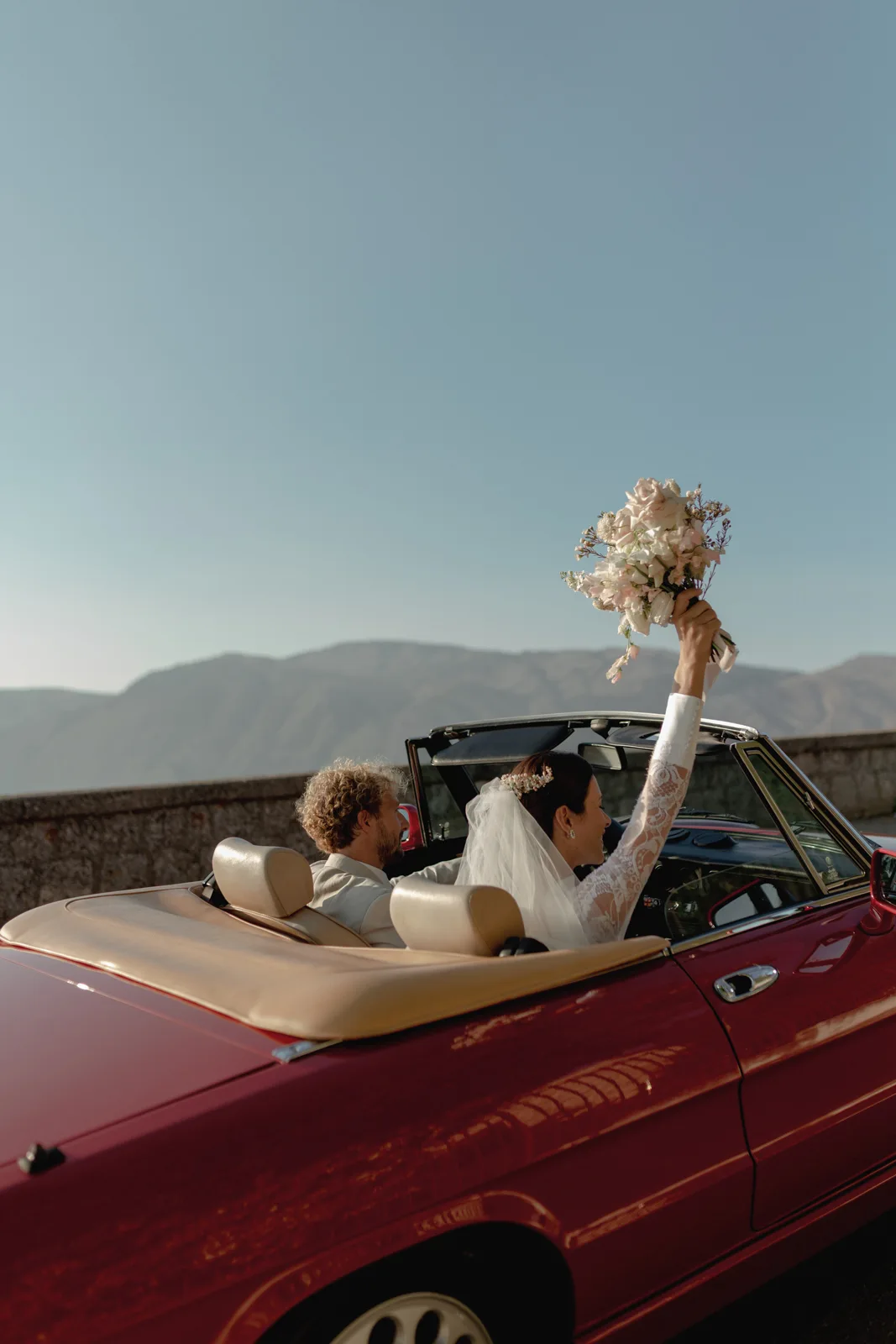 Home 19 A newlywed couple drives in a red convertible with mountains in the background. The bride, wearing a veil, holds a bouquet of flowers up joyfully while the groom drives. The sky is clear and sunny.