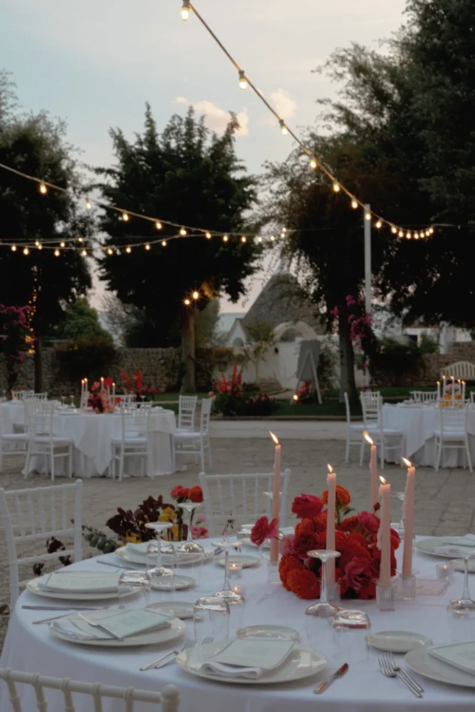 Outdoor evening event with round tables covered in white cloths, white chairs, and red floral centerpieces with lit candles. String lights hang above, and trees surround the elegant, festive setting.