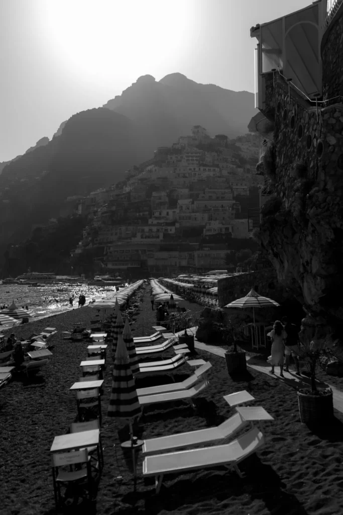 Home 14 Black and white photo of a beach with rows of empty lounge chairs and umbrellas, a rocky hillside covered with buildings in the background, and sunlight shining from behind the mountains.