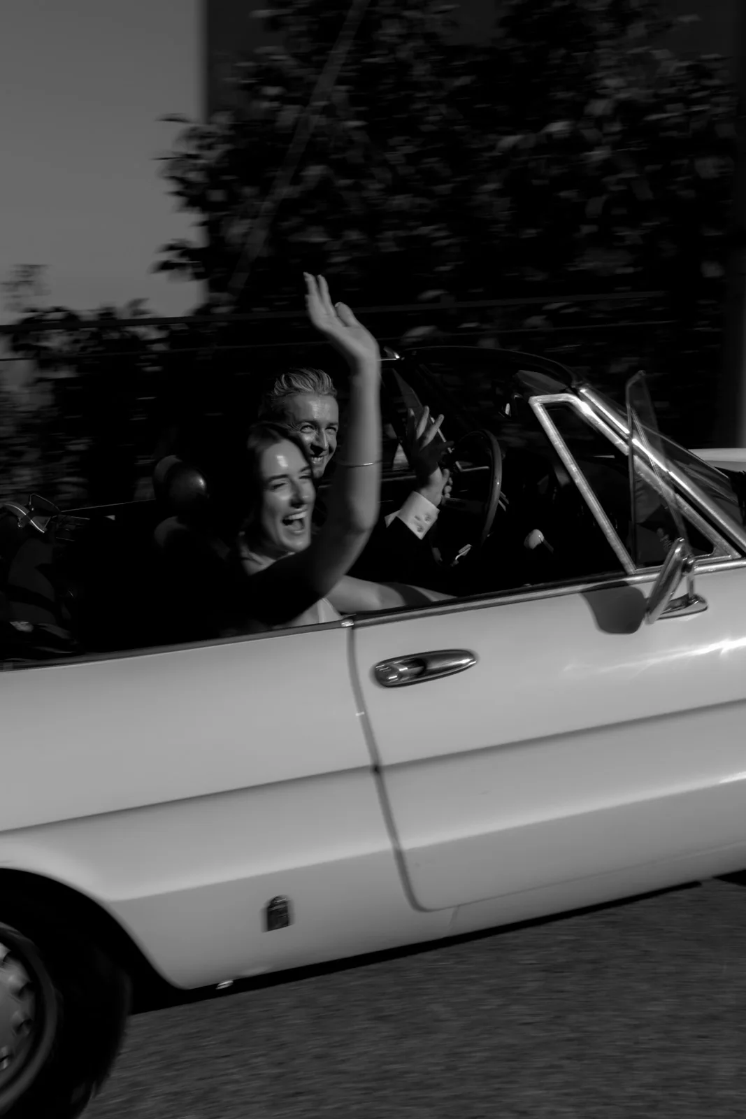 Home 40 A woman and a man smile and wave while driving a classic white convertible car. The image is black and white, capturing a sense of motion and joy as they enjoy the ride.