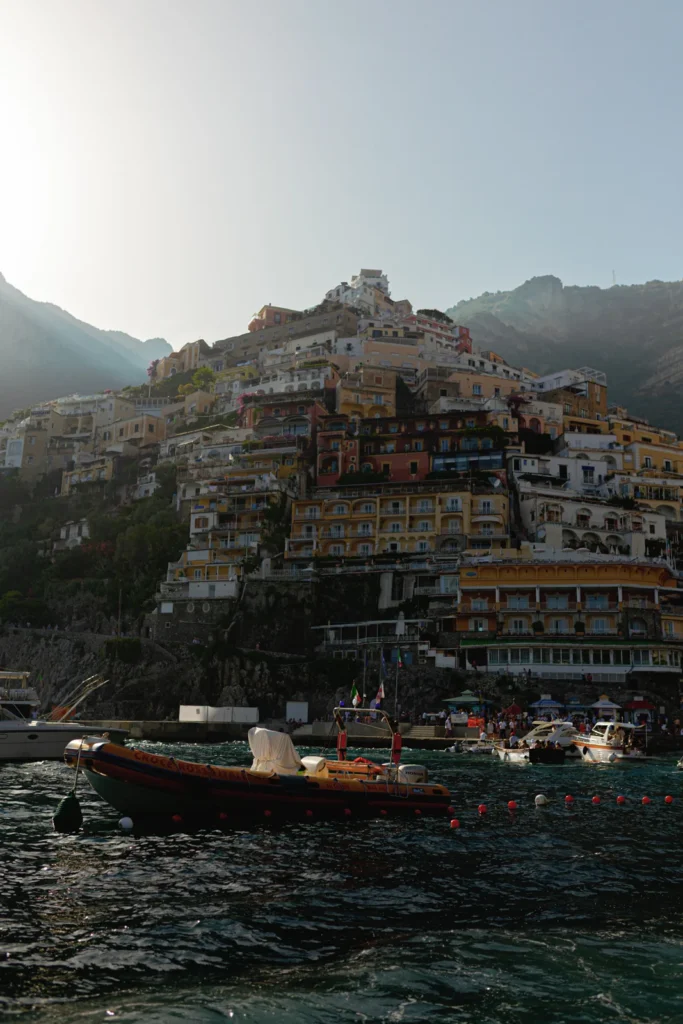 Colorful buildings stacked on a steep hillside in Positano, Italy, as seen from the water at sunset. Boats float on the blue sea in the foreground, with hazy mountains in the background.