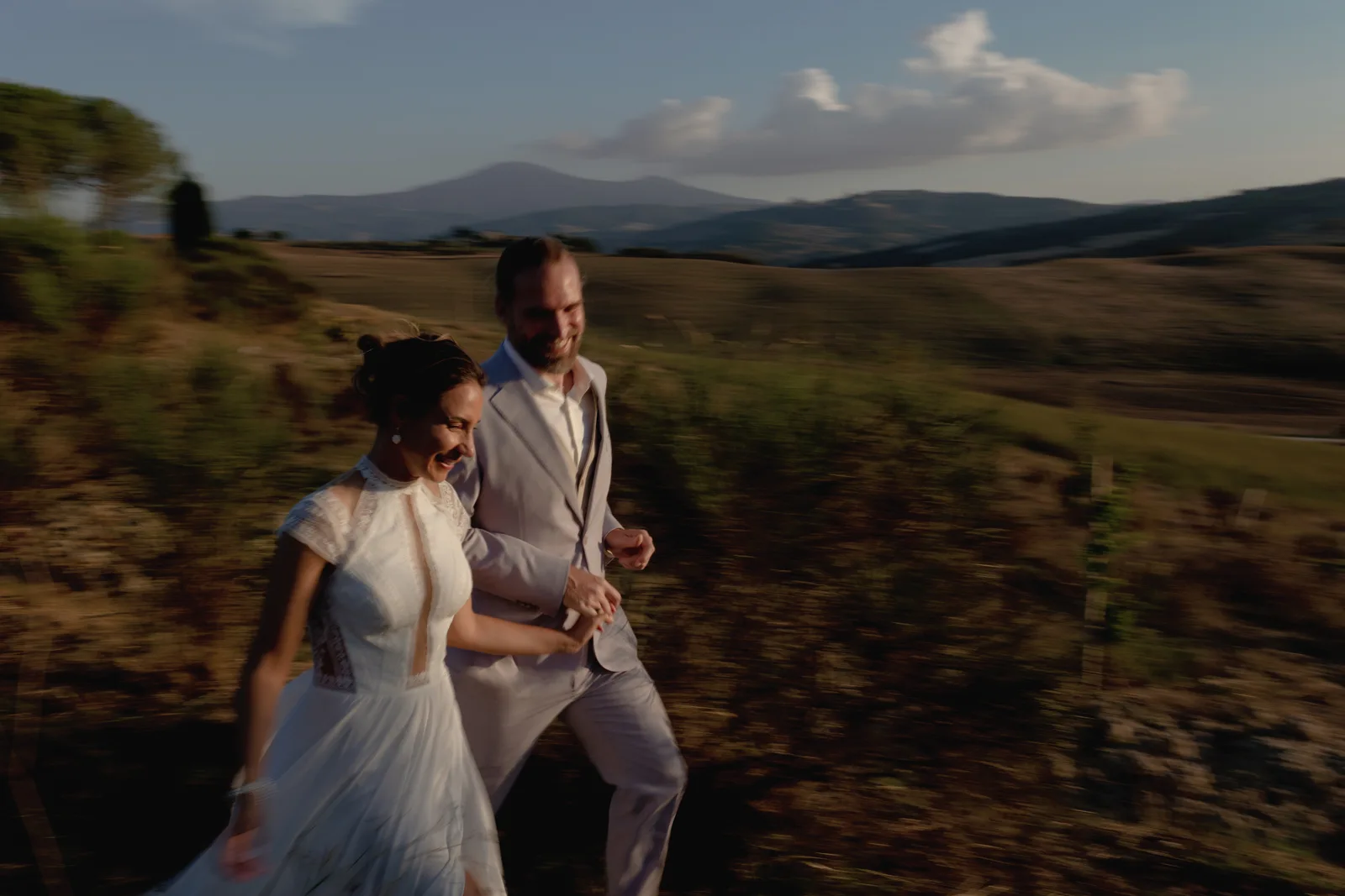 A bride and groom walk hand in hand outdoors, smiling, with hills and a mountain in the background under a blue sky with clouds, beautifully captured by a Tuscan wedding photographer in warm, natural light.