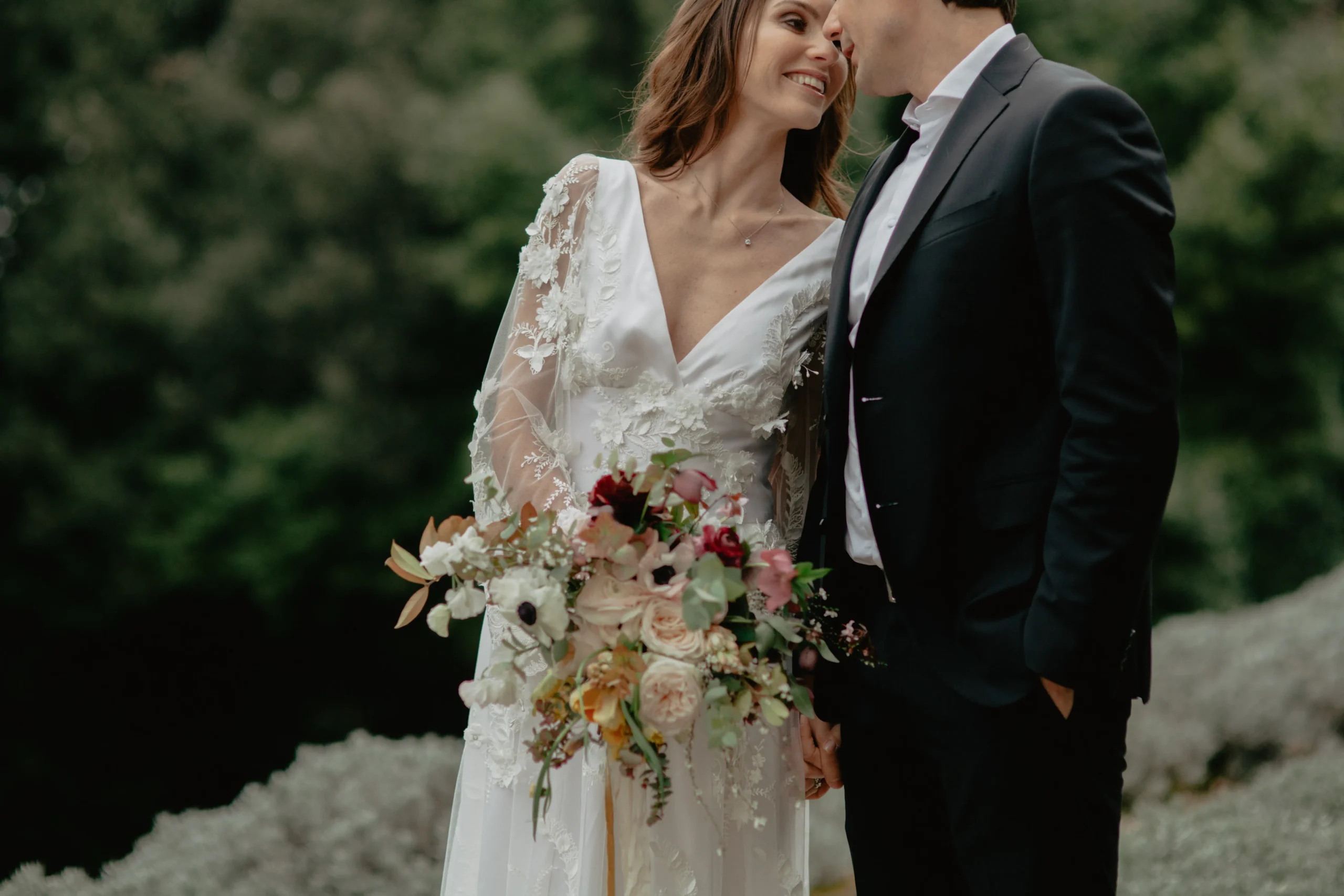 Kind Words 21 A bride in a white dress holding a bouquet smiles lovingly at a groom in a black suit as they lean close together outdoors, with greenery blurred in the background—a moment perfectly captured by an Italian wedding photographer.