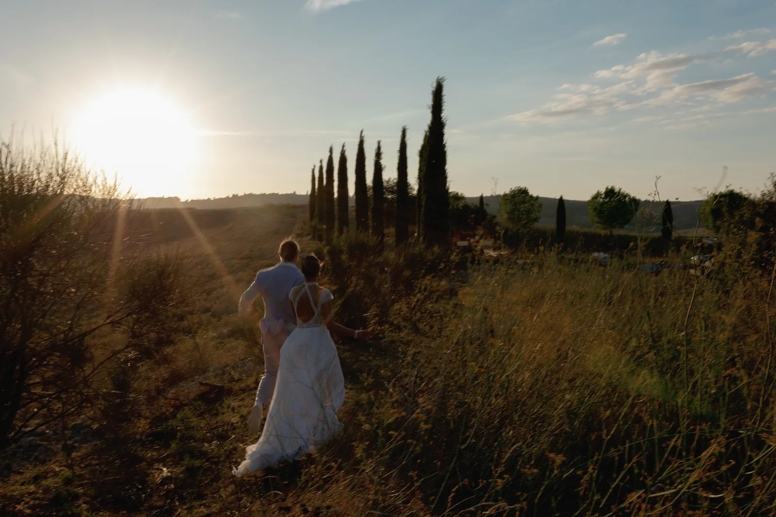 Home 22 A bride and groom walk hand in hand through tall grass at sunset, with the sun low in the sky and cypress trees lining the path in the background.