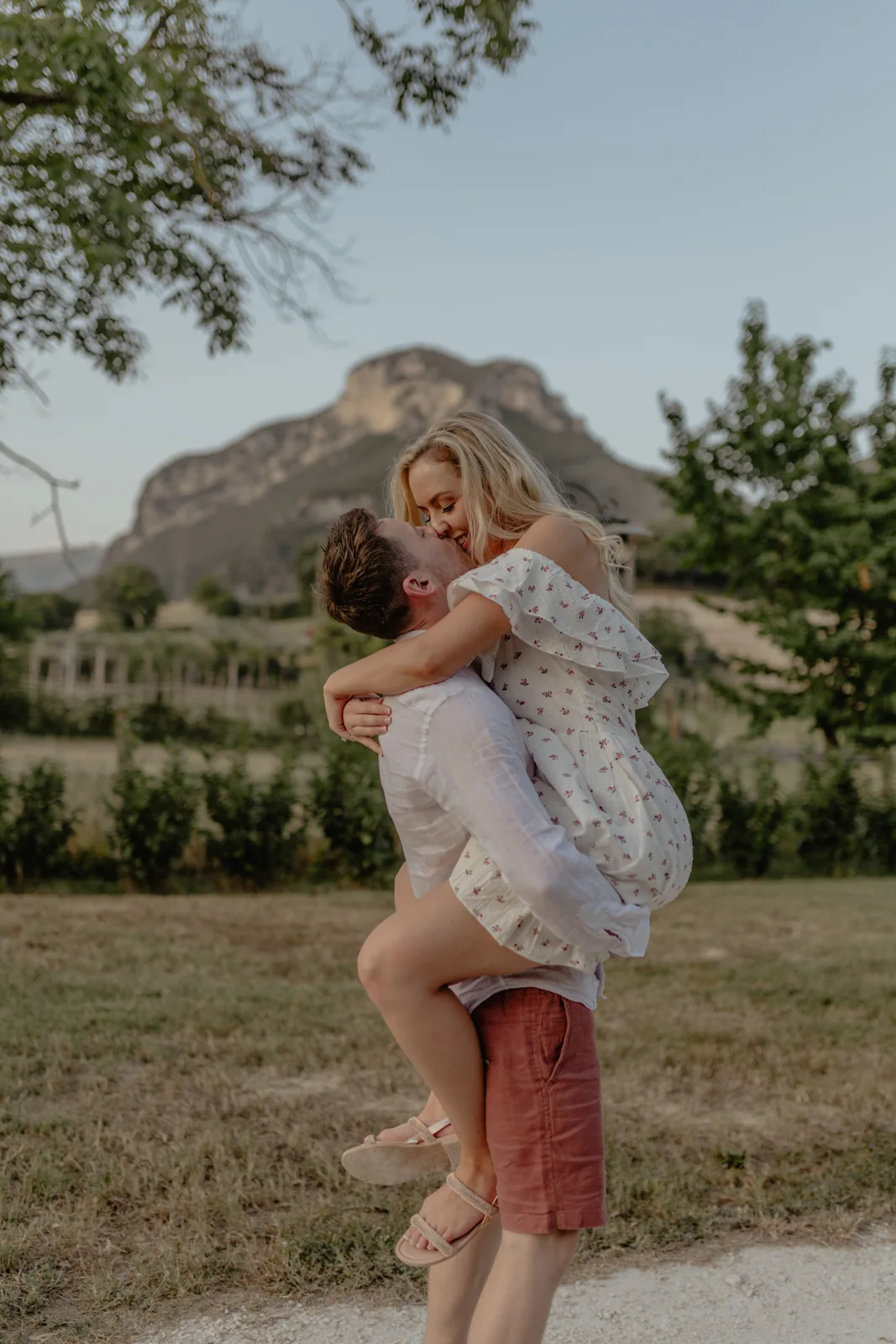 Home 20 A man lifts a woman in a white dress as she wraps her arms around his neck. They are outdoors in a grassy area, with trees and a mountain in the background, both smiling and looking happy.