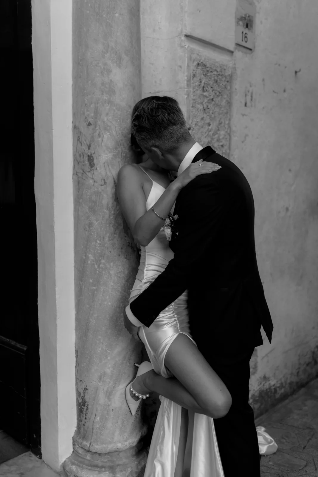 Home 25 A couple dressed in formal wedding attire passionately embrace against a column indoors. The woman wears a white gown and heels, while the man is in a dark suit. The intimate moment is captured in black and white.